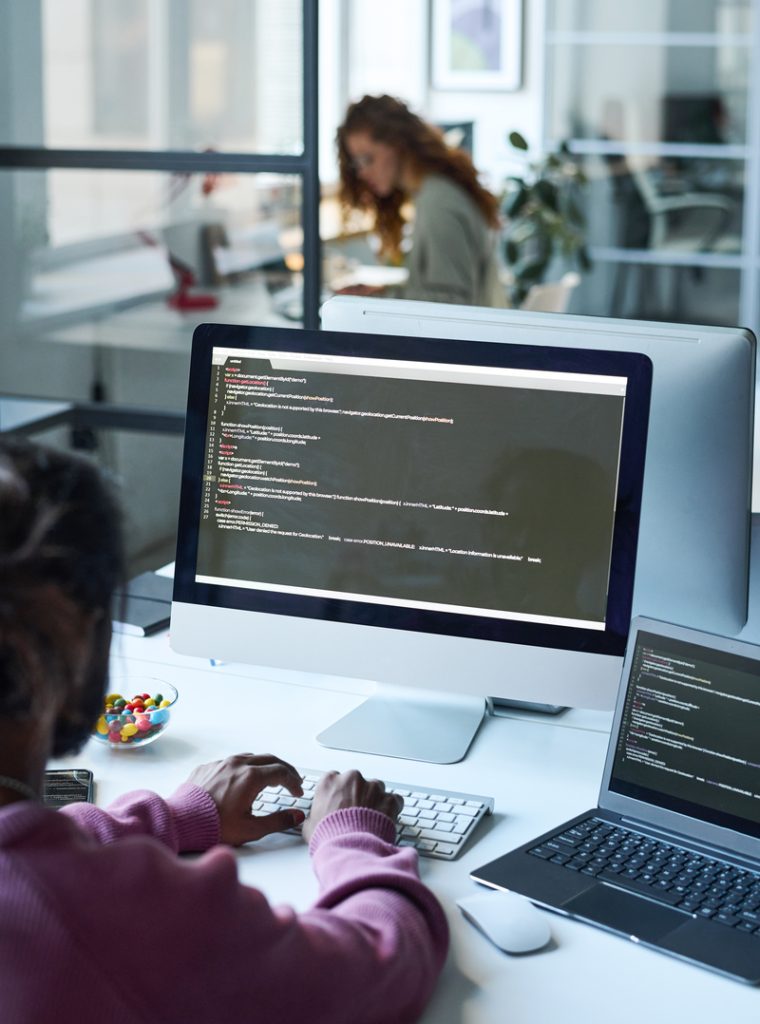 Rear view of young developer sitting at his workplace with computer monitor and typing codes for computer program